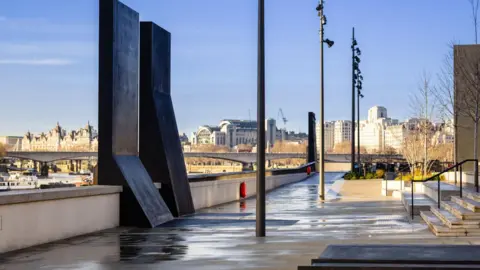 A smooth path and steps by a river, with a black oblong sculpture in the foreground and some concrete steps in view.