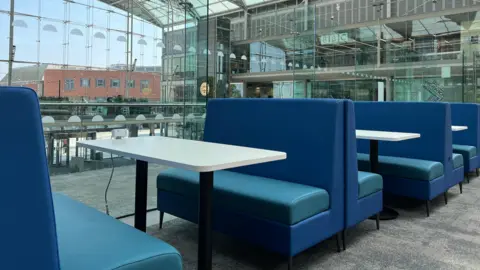 Norfolk County Council Blue sofa chairs line the first floor of the Norfolk and Norwich Millennium Library creating a new study space. The chairs are pushed up against the glass balcony with a table in between and plug sockets. The area looks over the rest of The Forum and outside the large glass building into the city of Norwich.
