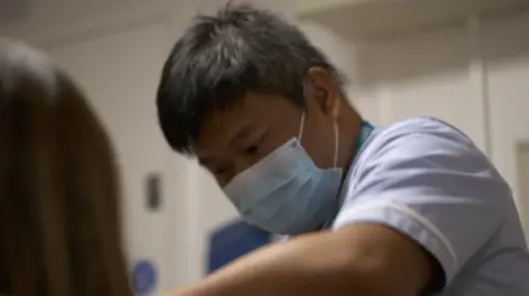 The image shows an Asian nurse, Tito Vicario, wearing a blue face mask and blue scrubs. He is leaning forward and taking the bloods of a female patient with long hair. He is in a white hospital ward.