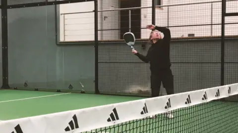 Padel Cornwall Tom Hutchins in a black sweatshirt and black jogging bottoms, he is positioned holding the padel racket up. He is in an indoor paddle court. The floor below him is green and the wall is grey. 