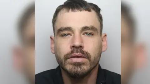 Police image of a man with dark hair and a short dark beard looking towards the camera wearing a black shirt 