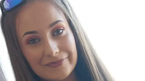 Chloe smiling at the camera. She has long straight light brown hair. She is wearing sunglasses on top of her head.