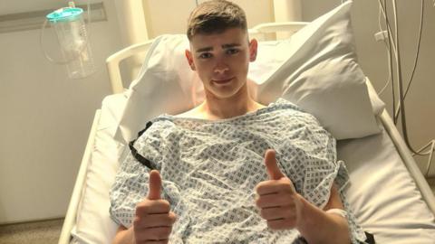 Jack Marshall in a hospital bed with his thumbs up