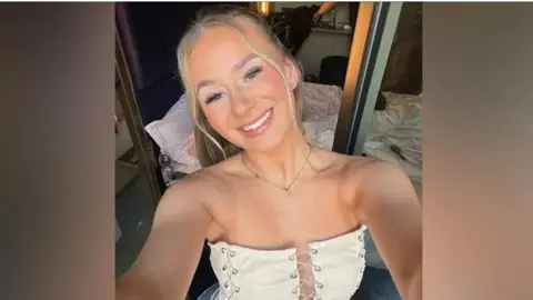 A woman with blonde hair takes a selfie. She's wearing a cream bodice style top and a silver necklace. She is smiling into the camera with a bed and mirrored wardrobe behind her