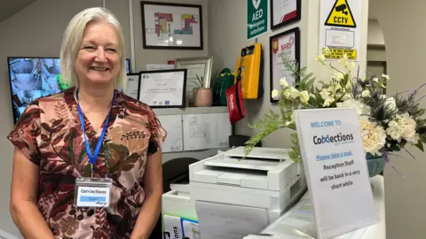 Clare Worden/BBC A woman stands behind a reception desk. She is wearing a patterned brown top. She has grey bobbed hair. She is wearing a blue lanyard which has her name on it. On the reception desk is a sign welcoming people and behind it are some flowers. 