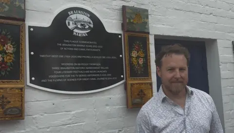 Martin Heath/ BBC An image of actor Samuel West standing next to a black and white commemorative plaque at Braunston Marina, surrounded by traditional "Roses and Castles" style painted canalware panels.