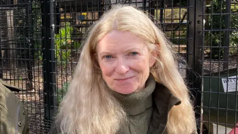 McKenna pictured with long blonde hair and wearing a dark green jumper and a dark green coat. She is stood in front of a black metal cage.