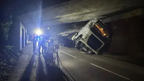 Essex County Fire and Rescue Service A white lorry is lying partially on its side under a bridge, at night-time. There are fire crews surrounding it.