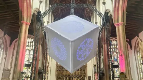 A grey cube, installed between two pillars in a church. Three sides of the cube are visible, and each had a geometric pattern on it. Purple light is shining through this. The pillars are lit up with pink lights. 