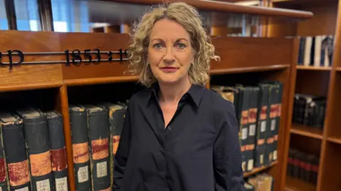 A woman wearing a black dress stands in front of old files 