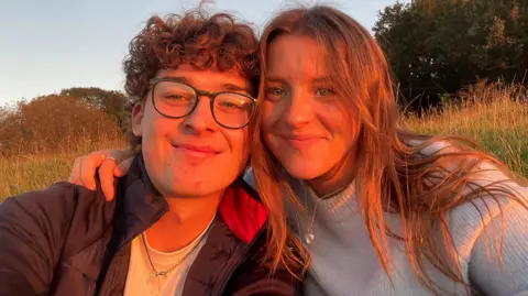 Ethereal Jewellery Hugh and Leah outside, with a golden sun glow on them, they are both looking at the camera. Hugh has on glasses, and has curly dark hair, Leah has long dark hair, is wearing a jumper, with a necklace round her neck.
