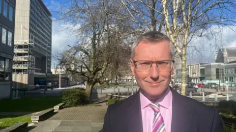 Patrick Nicholson, standing near Plymouth Civic Centre. He is a middle-aged man with short, greying hair and glasses, wearing a dark suit, a light pink shirt, and a striped tie.