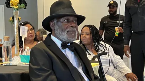 Guy Reid-Bailey looks off towards the right of the image, wearing a black suit with white shirt and black hat. In the background other party goers are visible