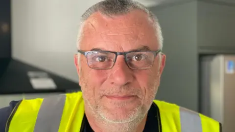 Stephen Huntley/BBC Kent Taylor is standing in a kitchen. He has short grey hair and stubble and is wearing black framed glasses, black T-shirt and a yellow Hi-Vis.
