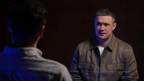 A man dressed in a white T-shirt and brown collared shirt in front of a black background sits talking to someone with their back to the camera.