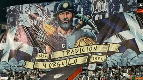 A large flag in a Mexican football stadium showing a Cornish miner holding a pasty and a pickaxe with two Cornish St Pirans flags either side of him. The flag shows the year 1892, when Pachuca were founded, as well as text that says 'tradicion y orgullo' - meaning tradition and pride in English. 