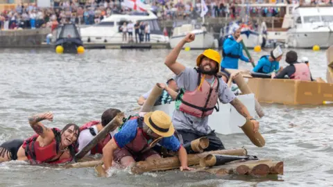 Bristol Harbour Festival An image of several people wearing hats, t-shirts and lifejackets clinging and sitting on three small boats made out of cardboard. A make-shift cardboard boat in the foreground appears to be sinking in the grey water. In the background can be seen a bridge lined with spectators.