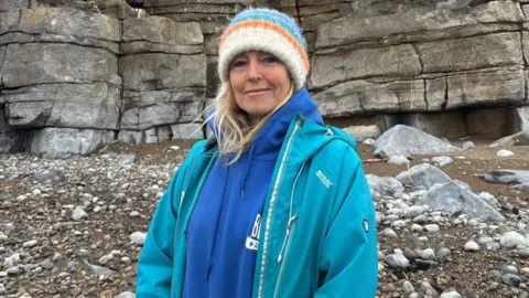 A woman with long blonde hair, wearing a white, orange and blue beanie hat, a blue hoodie and turquoise blue rain jacket. Behind her is a pebbly beach and cliffs. She is looking at the camera and smiling.