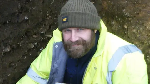 CAT Adam Wightman crouched in a Roman pit. He has a full beard and is wearing a woolly hat and a hi-vis yellow jacket over a blue fleece. Behind him can be seen earth and roots. 