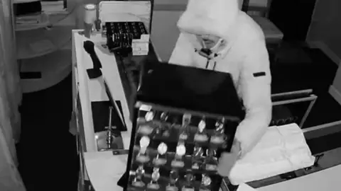 Cambridgeshire police Black and white image of a hooded man picking up a large display of wrist watches from a jewellery counter.