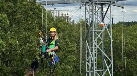 Birmingham Institute for Forest Research A woman in a harness hanging from a pylon next to a tree canopy. She has long blonde hair and wears a yellow hard hat, green vest and black t-shirt and trousers, with brown walking boots.