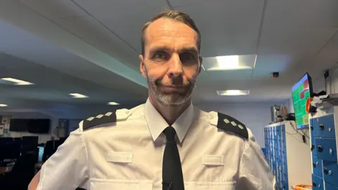 A chief inspector wearing a white shirt and a black tie standing in a police control room