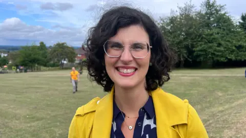 Layla has medium length dark hair and wears hexagonal glasses, a yellow coat, and a black and white patterned collared shirt. She is standing in a park.