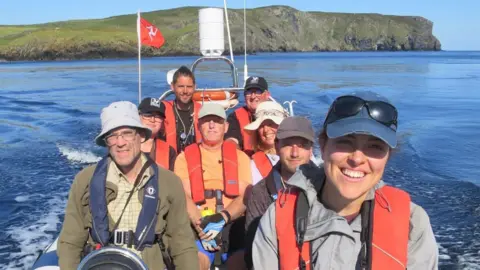 MWT Eight people sitting on a small boat. They are all wear orange lifejackets, and there is a Manx flag flying at the back of the boat, and the Calf of Man in the background.