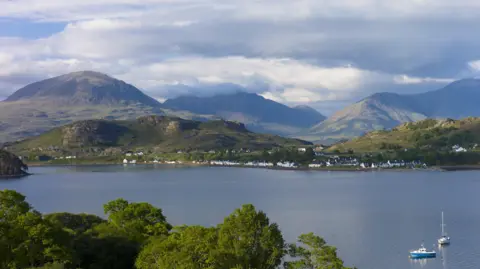 The loch is large and mountains rise from the shore line. There is a row of white-walled houses along the edge of the loch and two boats on the water.