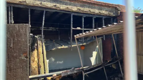 Shariqua Ahmed/ BBC A fire-damaged outbuilding with a corrugated iron roof that has partially collapsed. 