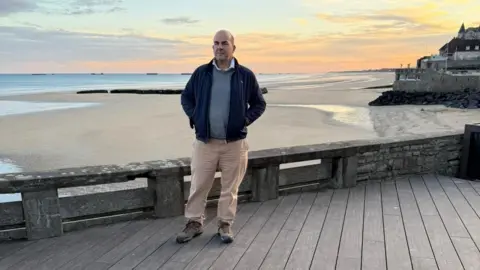 A bald man wearing a navy blue jacket, grey humper and beige trousers stands on a wooden platform with a beach behind him