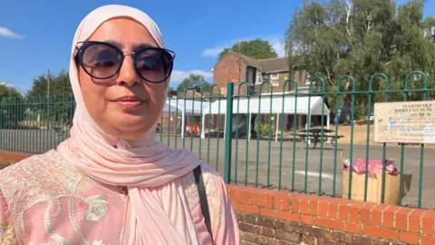Louise Parry/BBC Khtija looks at the camera, with a gentle smile. She wears sunglasses and a pink hijab, and a pink embroidered dress. She is standing outside a toddlers' play area, in front of a brick wall with metal railing on top of it.