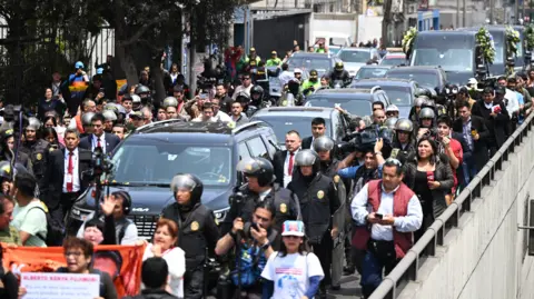 Getty Images De lijkwagen met het lichaam van voormalig president Albert Fujimori wordt gevolgd door beveiliging, politie, journalisten en cameramannen terwijl een konvooi zwarte voertuigen op weg is naar zijn uitvaartdienst.