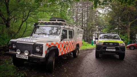 LOMOND MRT An image of two offroad vehicles in the Lomond Mountain Rescue livery parked up on a woodland path on the West Highland Way. 