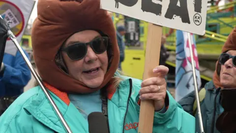 A woman wearing a turquoise coat while holding a placard. She is wearing a brown hat and black sunglasses