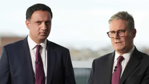 Two men stand side-by-side posing for a photograph. They are both wearing dark suits, white shirts with ties. 