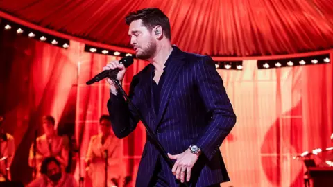 Getty Images Michael Bublé singing into a microphone with red lighting. He is wearing a dark blue pin-stripe suit and watch.