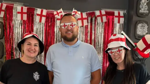 Holy Trinity Tattoo Studio Standish England football fans Amanda (left) Sean Carrington (centre) and Jamie Taylor (right) all dressed up with England red and white glasses and bucket hats. They are all smiling and standing in front of a shiny red and silver backdrop with England flags. 
