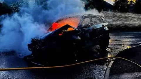 A black car on a road with bright orange flames inside it and white smoke emanating. A yellow hose can be seen crossing the road in front of it. It is sunset. 