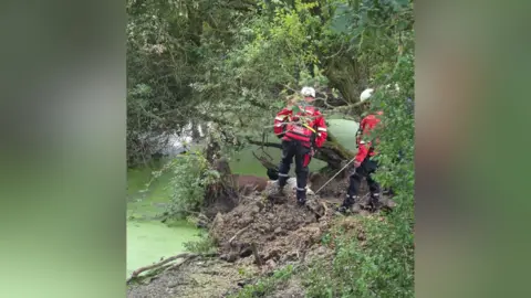 Dorset & Wiltshire Fire and Rescue Service Two firefighters seen by the pond where the bull was stuck. It is a cloudy day.
