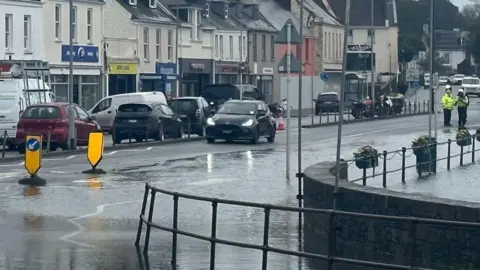 BBC A rain-soaked town street with standing water flooding the roadside, cars driving through wet conditions, and traffic officers visible in the distance near a row of shops and houses under grey skies.
