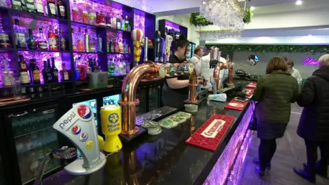 Workers behind a brightly-lit bar with gold-coloured pumps, red beer mats and drinks displayed on the rear shelf