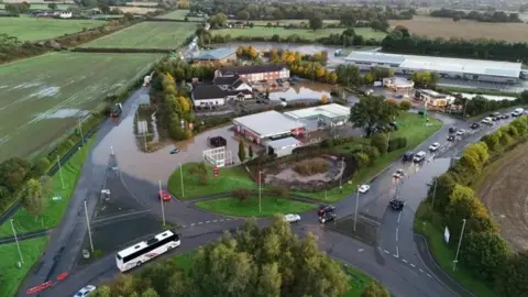 Carl Bishop A drone view of buildings and a petrol station surrounded by floodwater. The buildings are adjacent to a roundabout. There is one car submerged in flood water, and a queue of traffic building to the right hand side of the site