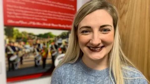 A woman with long fair hair is smiling at the camera. She is wearing a grey jumper and is standing in front of a picture of a marching band.
