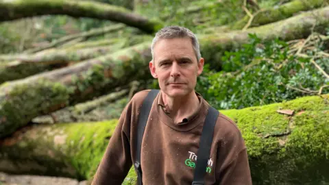 A man stands in front of a felled tree. The trunk is covered in green moss. The man is wearing a brown sweatshirt that says Trebah Garden on it.