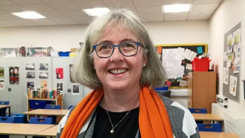 Victoria Rowe Victoria Rowe is a white woman in middle age. She has grey bobbed hair with a fringe. She wears a black top with a grey and white jacket and an orange scarf draped around her neck, as well as a silver necklace with a pearl pendant. She has blue-grey framed classes and is smiling. She is standing in a classroom with a few rows of desks and chairs behind her, cupboards with lots of pictures stuck to them and two pinboards on the walls.