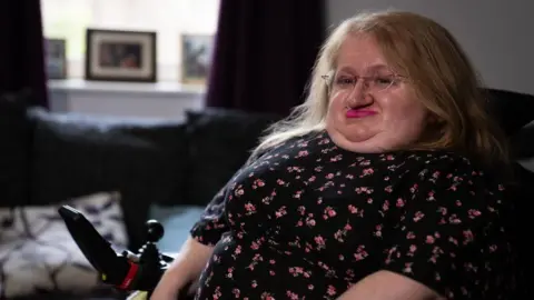 BBC News A woman with long blonde hair, glasses, and pink lipstick sits in a wheelchair. The woman is wearing a dark dress with flowers on it. 