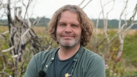Shaun Whitmore/BBC Andrew Waddison standing outside on the Wild Ken Hill reserve. He is looking directly at the camera and smiling and has long brunette hair. He is wearing a green T-shirt with a Wild Ken Hill logo on it. He is also wearing a green button-up shirt which is undone over the top of it.