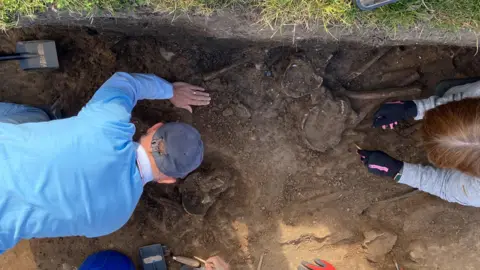 An aerial view of two people working in an archaeological dig with the back of one man taking up a third of the picture and the hands of a woman working around two skulls.