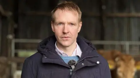 Peter Gallagher is looking directly at the camera. He is wearing a navy coat and has a blue jumper underneath and a pink and white striped shirt. He has blonde hair.